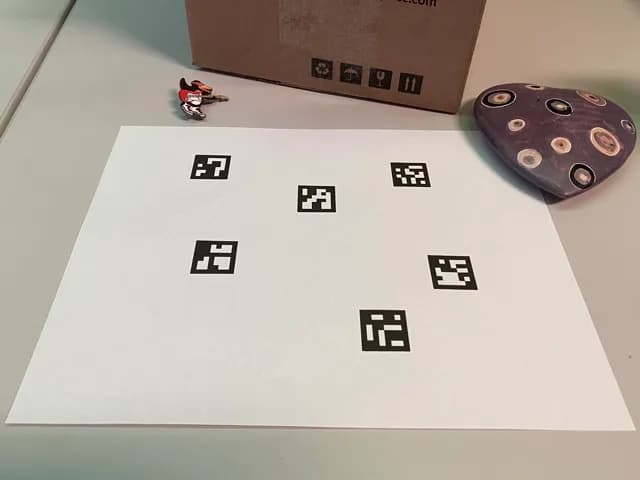 Hoja de papel con seis marcadores ArUco sobre una mesa, junto a una caja y un objeto de cerámica